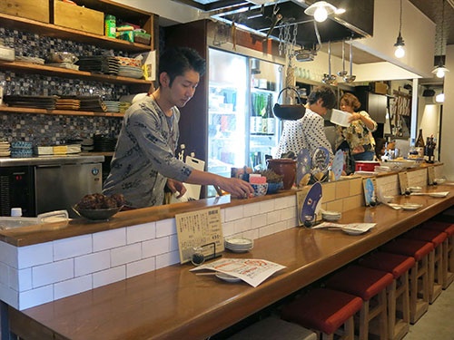 東京・笹塚に今年5月オープンした「炭火と酒 さささのさ」。店主は楽コーポレーションOBの片桐英二氏（左）。常時約20種の日本酒を揃える。出店場所は値段がある程度高くてもいいものを食べたいお客が多いエリアと判断し、鮮魚の炭火焼きを看板料理に据えた。店舗面積は15坪で2フロアに分かれ、2階には10人が座れて宴会利用がしやすい掘りごたつ席がある
